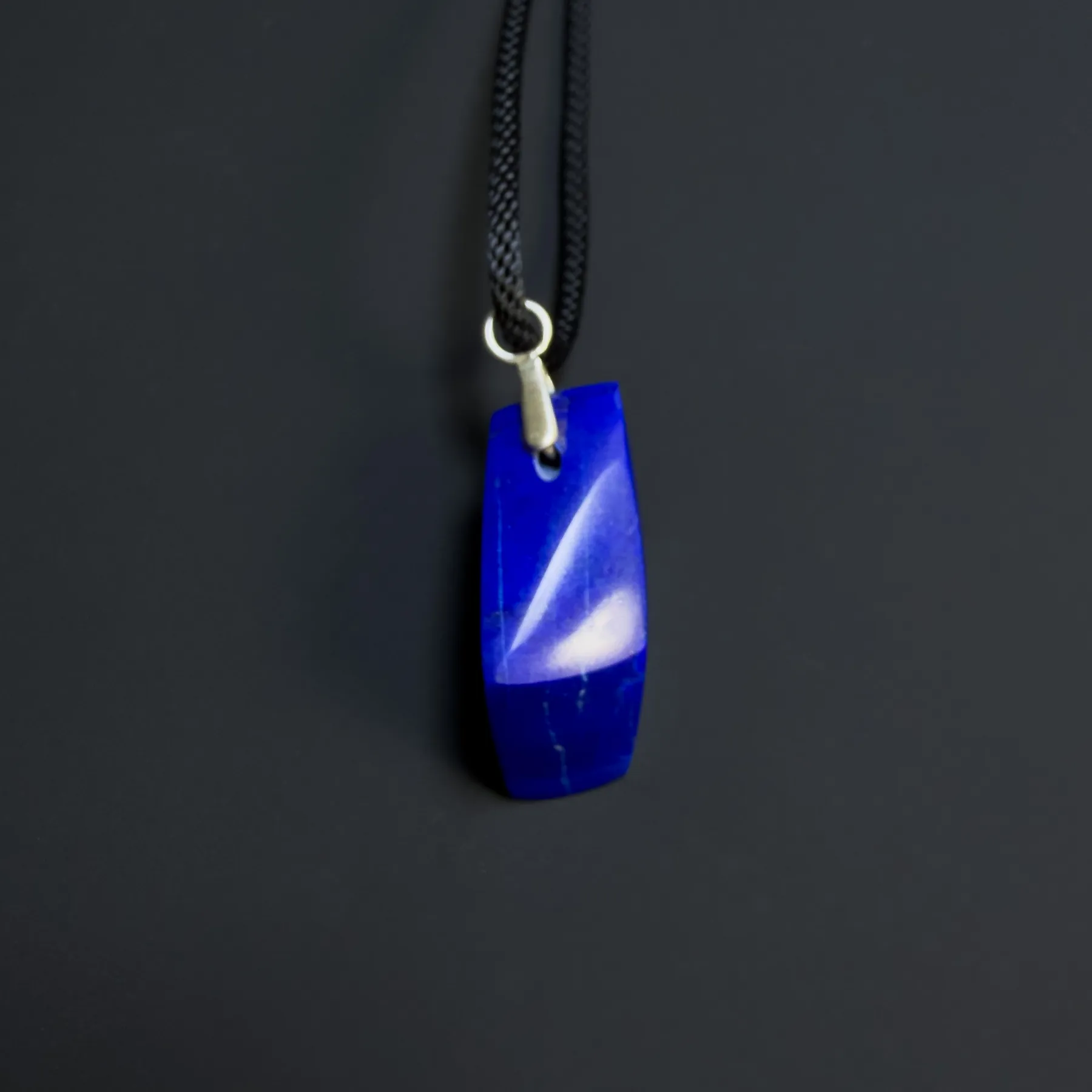 Narrow edge of the blue lapis pendant below a silver bail threaded onto black cord
